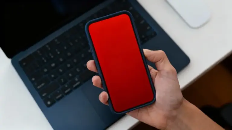 Una mano tiene uno smartphone con schermo rosso davanti a un laptop